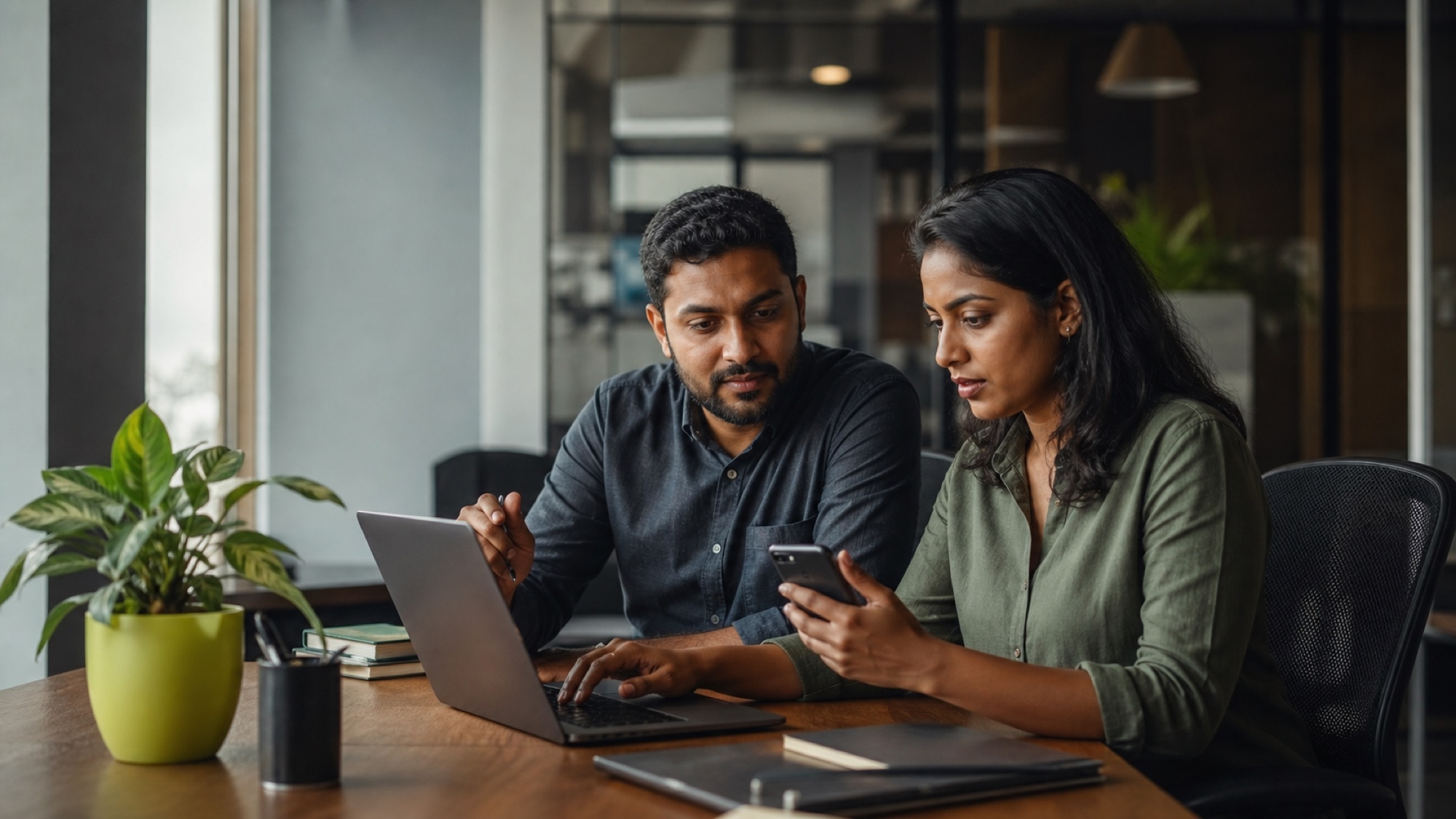 Two customer operations teammates compare a message on a phone with related information on a laptop.